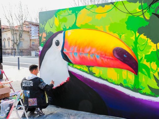 L'Art prend l'Air Perpignan 2026 Un artiste peint un toucan coloré sur un mur, entouré de verdure.