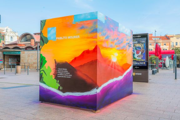 L'Art prend l'Air Perpignan 2026 Cube coloré avec un paysage vibrant au coucher de soleil et des éléments graphiques.